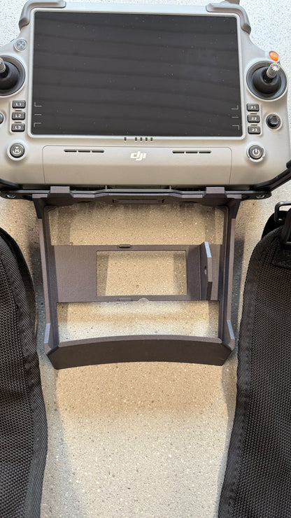 Ein DJI-Drohnen-Controller, ausgestattet mit einer schwarzen CIELOTEC RC Plus 2-Distanzhalterung – SDR- & Lautsprecher-Version (3D-Druck PA12), die für die Verwendung mit dem DJI RC Plus 2-Sendertragegurt (M4E, M4T) ist darunter angebracht und ruht auf einer gesprenkelten Oberfläche, wobei auf beiden Seiten schwarze Stoffriemen und ein DJI-Sendertragegurt zu sehen sind.
