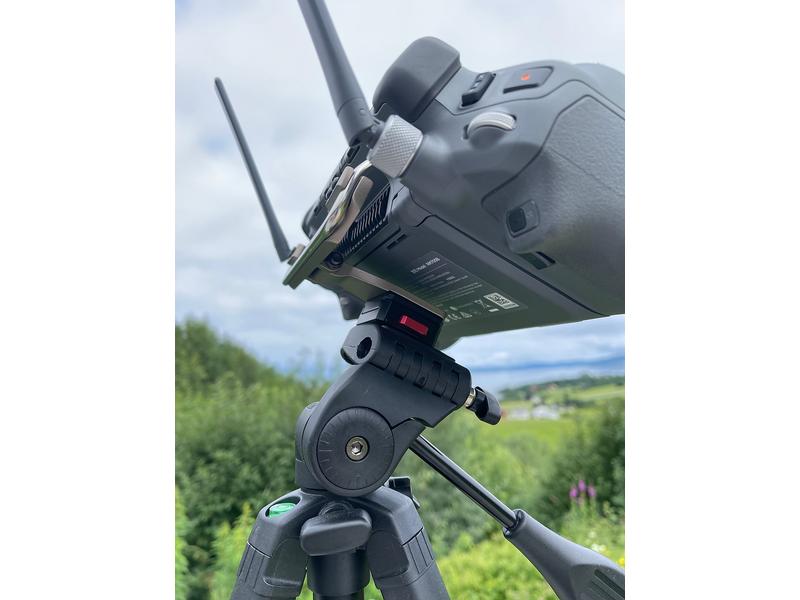 Nahaufnahme einer LifThor Stativhalterung für die DJI RC Pro Plus-Fernbedienung, montiert auf einem Aluminiumständer. Das Gerät mit Doppelantenne und mehreren Tasten ist vor üppigem Grün und einem bewölkten Himmel zu sehen.